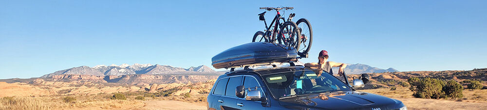 Woman standing outside an SUV with a cargo box and bikes mounted on the roof, scenic buttes in the background.