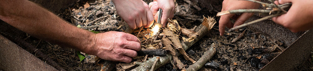 Three sets of hands adding tinder and using a fire starter in a fire pit.