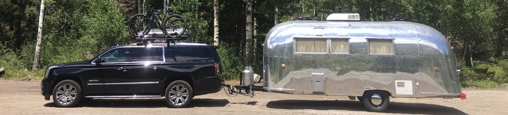 Black Jeep with bikes and cargo box on roof pulling Airstream camper with forest in background.